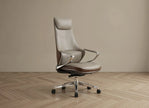 A modern high-back office chair upholstered in light taupe leather with dark brown leather trim, featuring chrome armrests and a five-star metal base with black casters, set against a minimalist beige wall and light wood herringbone floor.
