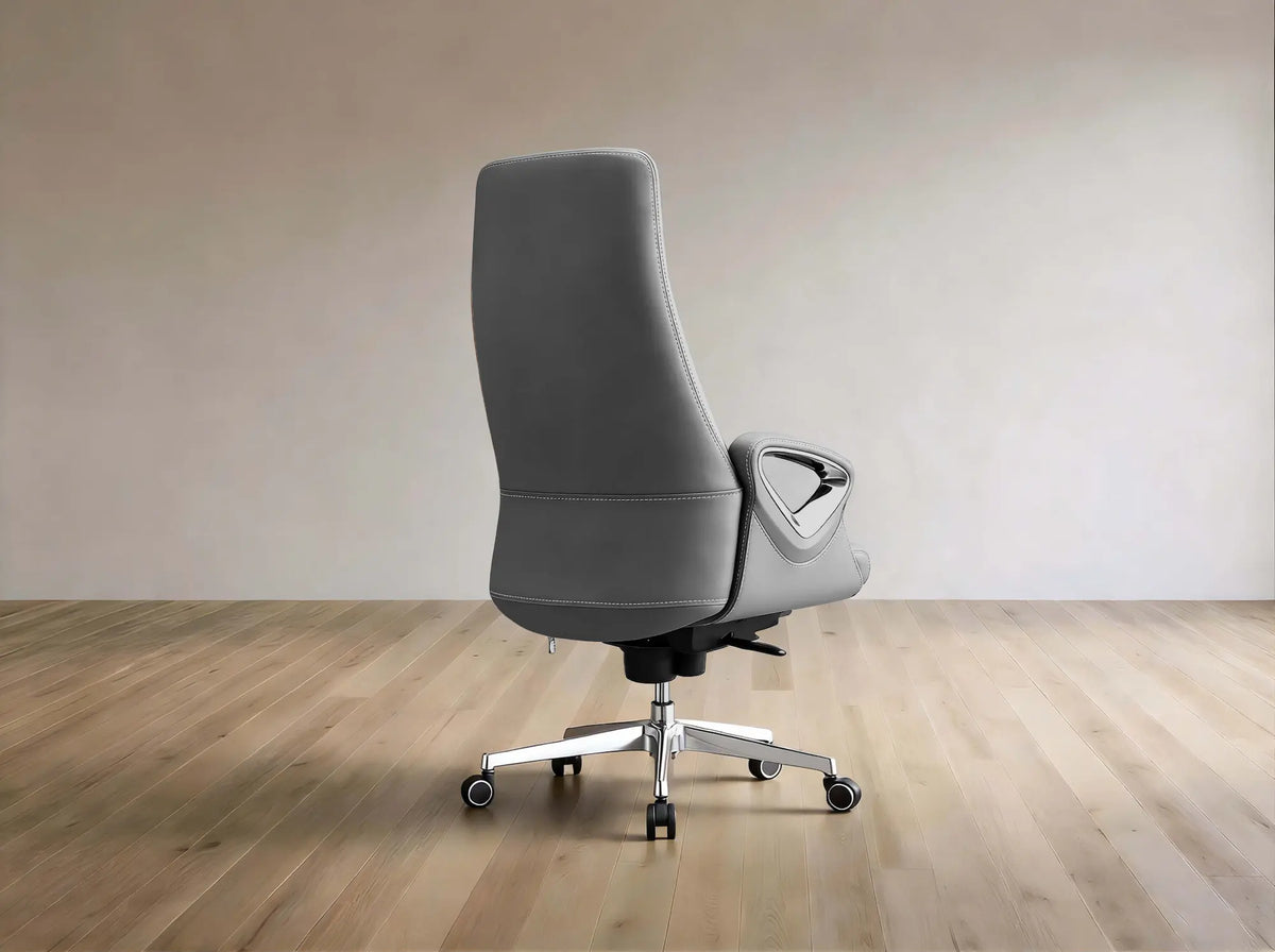 A modern gray upholstered office chair with a high back, curved armrests, and a chrome five-star wheeled base, shown in a minimalist room with light wood flooring and a beige wall.