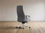 A modern gray upholstered office chair with a high back, curved armrests, and a chrome five-star wheeled base, shown in a minimalist room with light wood flooring and a beige wall.