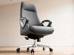 A modern black leather office chair with a high back, chrome five-star base, and adjustable armrests, shown in a minimalist room setting with a beige wall and light wood floor.