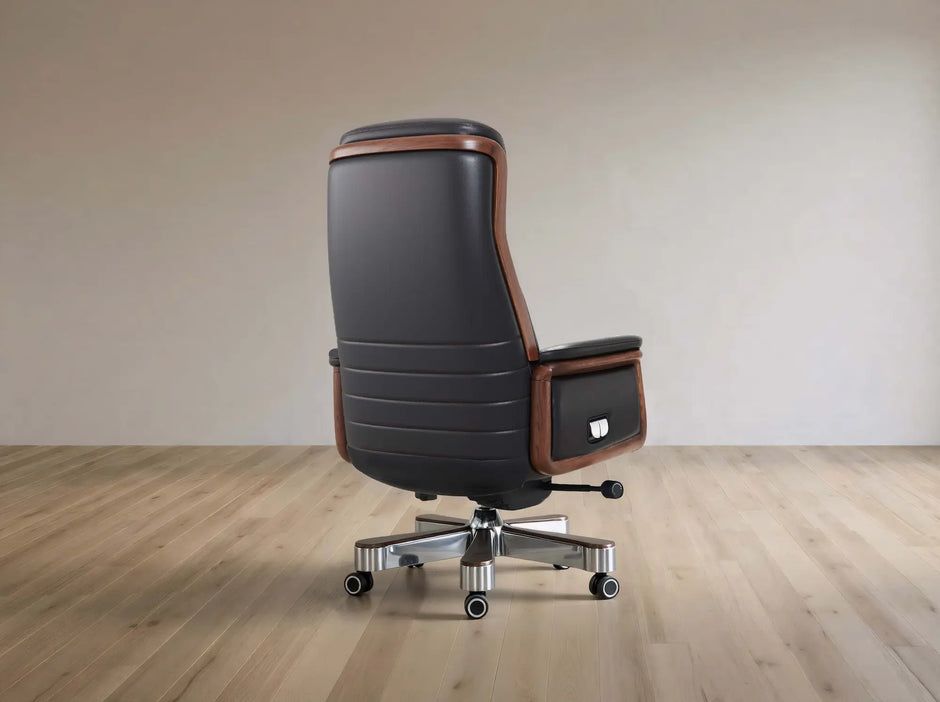 A high-back office chair with black leather upholstery and brown wood trim, mounted on a chrome five-star wheeled base, shown in a minimalist room with light wood flooring and a beige wall.