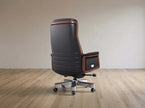 A high-back office chair with black leather upholstery and brown wood trim, mounted on a chrome five-star wheeled base, shown in a minimalist room with light wood flooring and a beige wall.