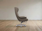 A modern high-back lounge chair upholstered in taupe leather, shown from the side on a light-colored wooden floor against a neutral beige wall.