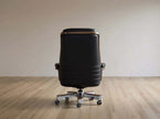 A black leather office chair with a high back, silver five-star base, and wood trim, shown from the rear on a light wood floor against a neutral beige wall.
