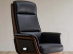 A high-back black leather office chair with dark brown wood trim and a small side control switch, photographed against a plain light beige wall.