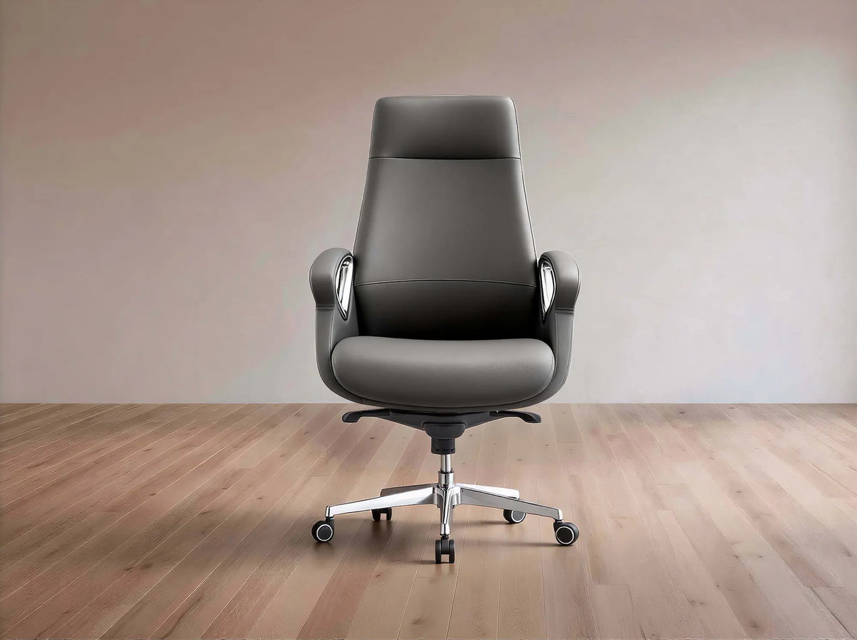 A dark gray leather office chair with a high back, silver armrest supports, and a chrome five-star wheeled base, set against a minimalist beige wall and light wood floor.