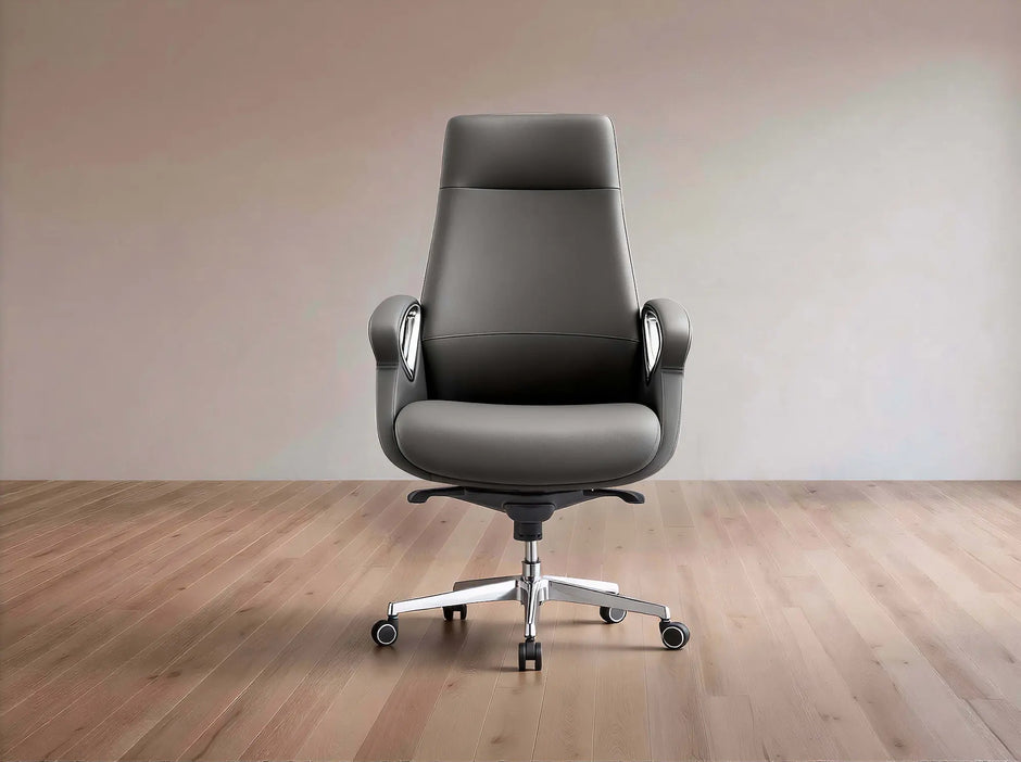 A dark gray leather office chair with a high back, silver armrest supports, and a chrome five-star wheeled base, set against a minimalist beige wall and light wood floor.