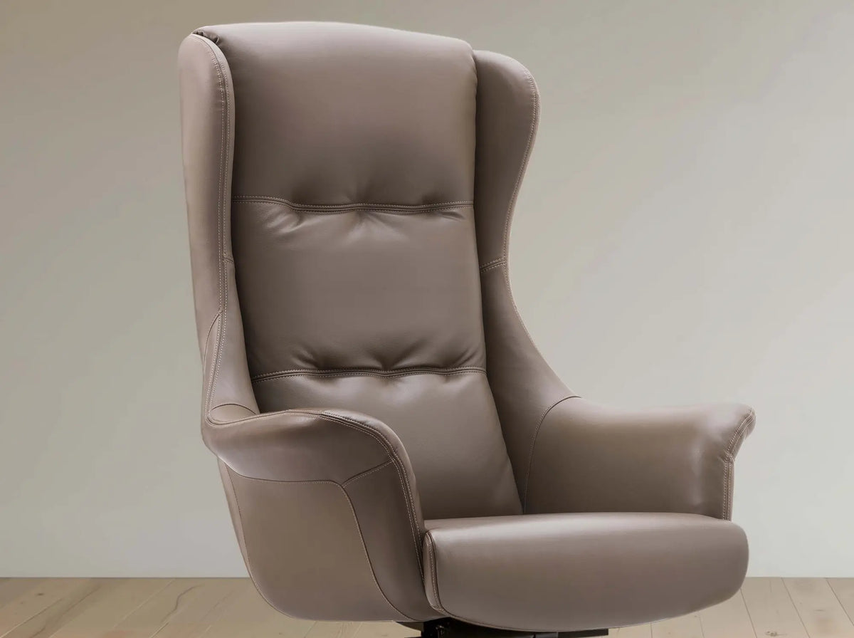 A high-back wingback office chair upholstered in taupe leather, shown from a three-quarter angle against a neutral beige wall and light wood floor.