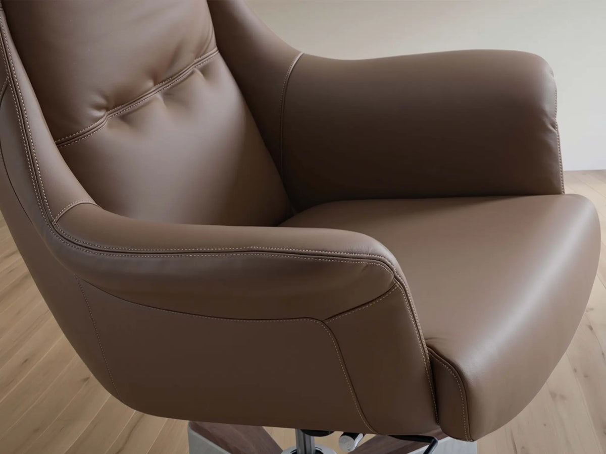 A close-up of a modern armchair upholstered in smooth taupe-brown leather, showing the seat, armrests, and tufted backrest, set against a light wood floor and neutral wall.