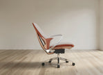 A modern ergonomic office chair with a curved backrest, upholstered in a terracotta orange and beige leather-like material, featuring a chrome metal armrest and a five-star black wheeled base, set against a minimalist beige wall and light wood floor.