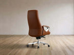 A modern brown leather office chair with a high back and chrome five-star base, shown from the side against a minimalist beige wall and light wood floor.
