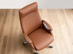 A modern brown faux-leather office chair with chrome armrest accents and a five-star wheeled base, shown from an elevated angle on a light wood plank floor against a white wall.