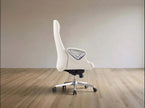 A modern white gaming chair with a high backrest, silver armrest frame, and chrome five-star wheeled base, shown in a minimalist room with light wood flooring and a beige wall.