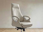 A high-back office chair upholstered in light beige leather with curved armrests and chrome accents, shown on a black wheeled base against a neutral beige wall and light wood floor.