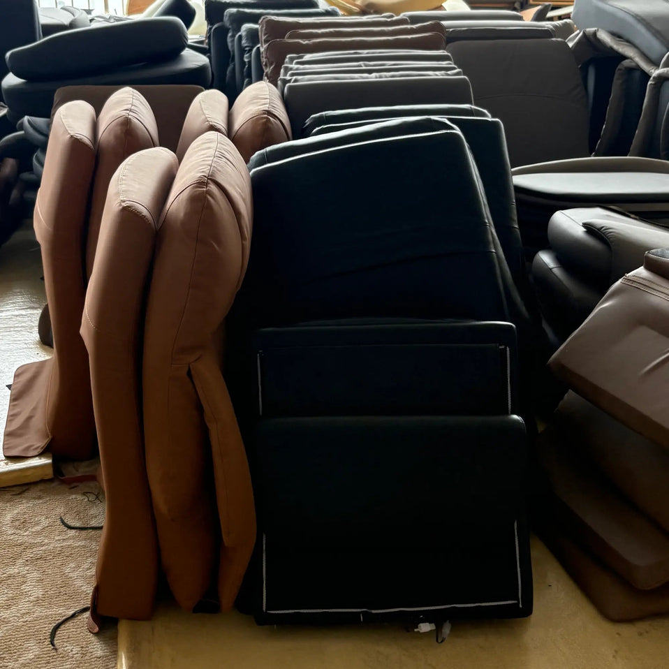 A warehouse or showroom floor filled with stacks of upholstered chair and sofa components, including tan cushions, black seat backs, and dark chair frames, near large windows.
