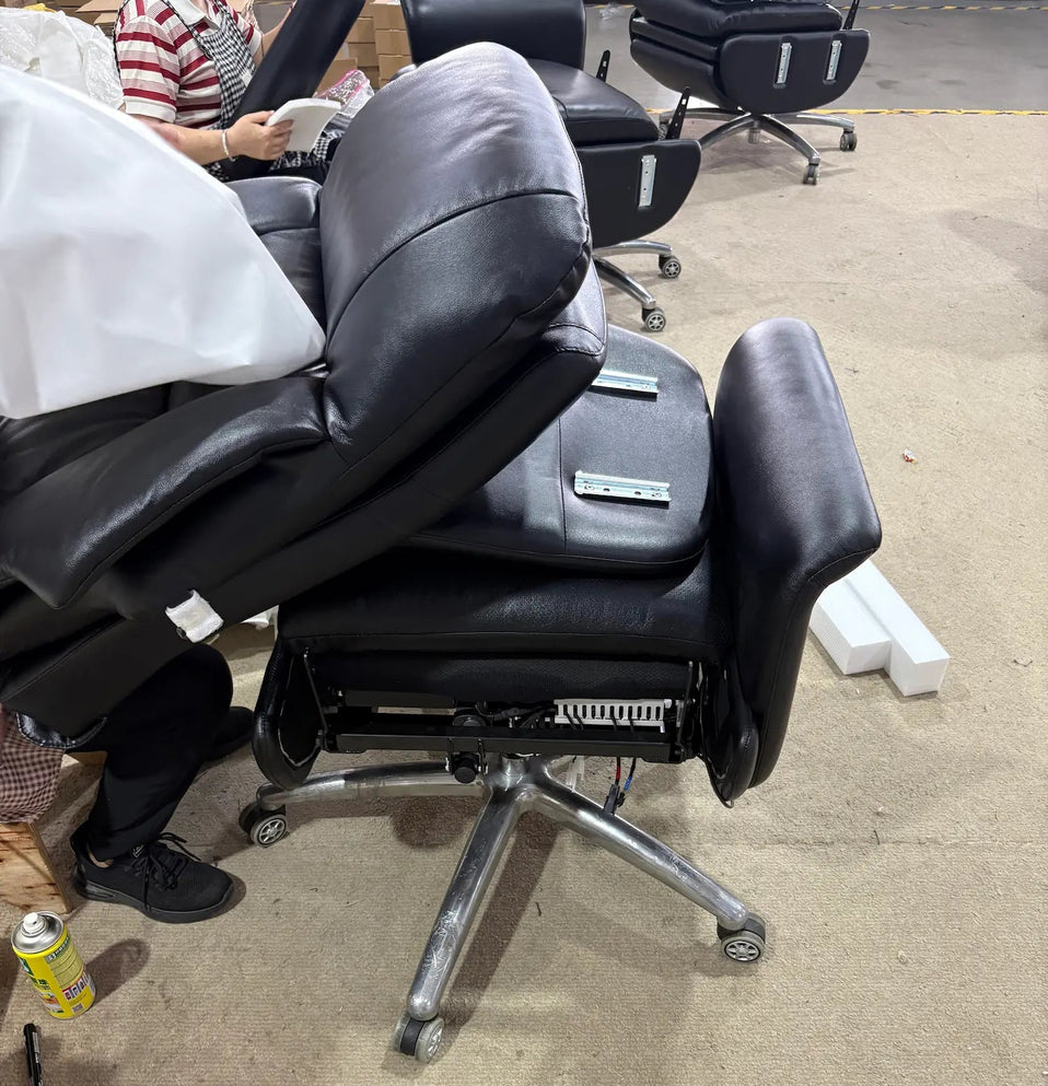 A black leather office chair with a reclining backrest and chrome five-star base, photographed in a warehouse or showroom setting on a concrete floor. A person is partially visible behind the chair, and other chairs and tools are visible in the background.