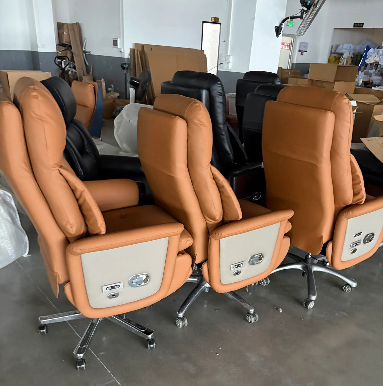 Three tan leather office chairs with cream side panels and chrome rolling bases, photographed in a warehouse-like setting with other chairs and cardboard boxes in the background.