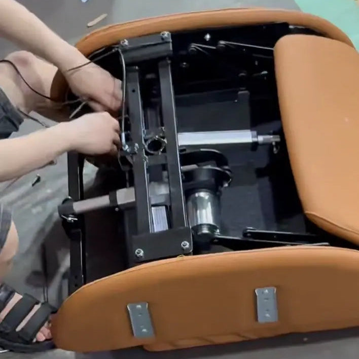 A person assembling a modern leather chair seat with a black metal frame mechanism, featuring tan leather upholstery and silver hardware, set on a gray floor.