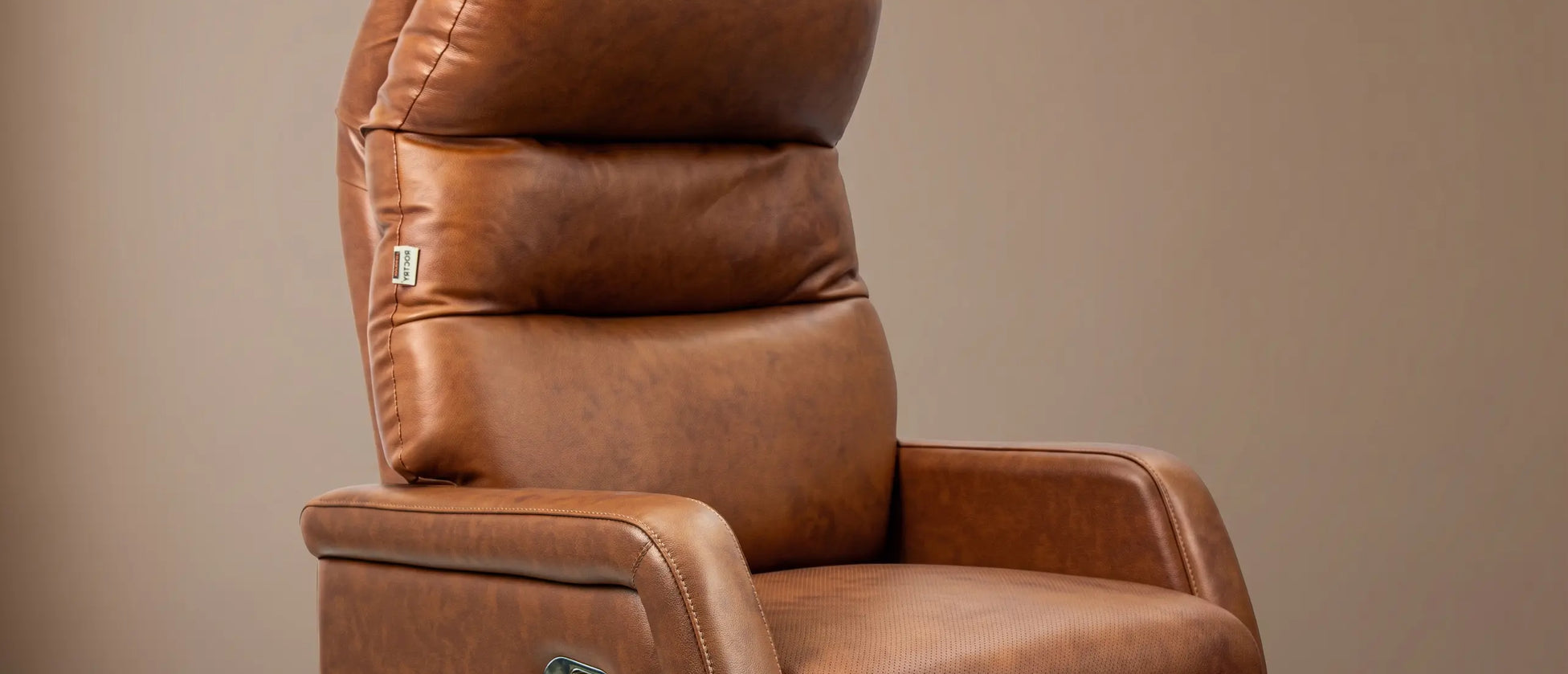 A brown leather recliner chair with a high backrest, shown in a studio setting against a neutral beige wall and dark wood floor.